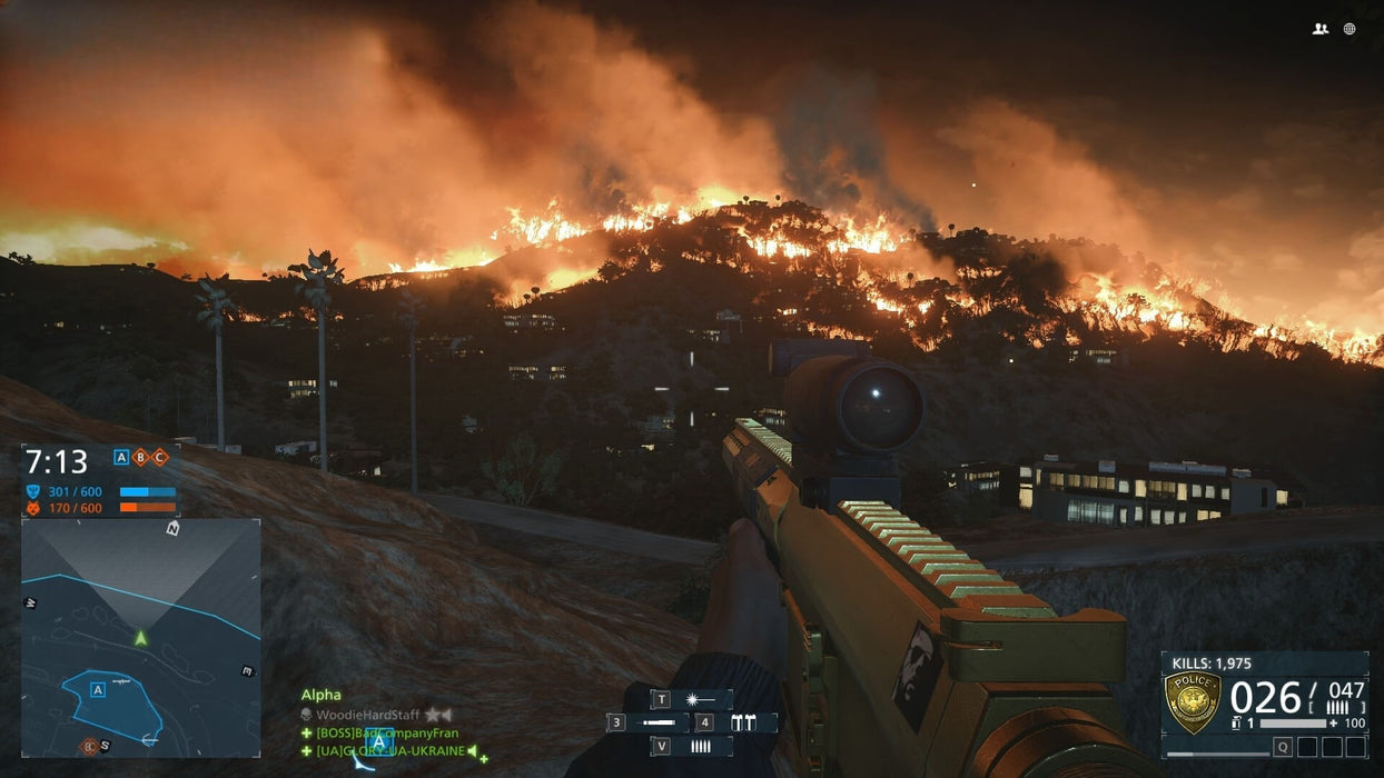 A first-person shooter perspective in Battlefield Hardline, showing a sniper rifle aimed at a massive wildfire engulfing a hill at night. The orange flames contrast with the dark sky as smoke billows upwards, creating an intense battlefield atmosphere