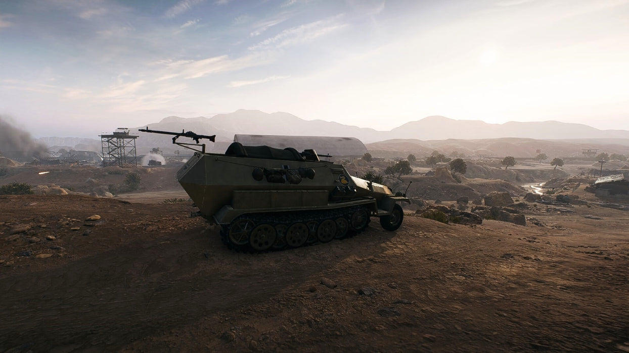 A World War II-era armored half-track vehicle parked in a dusty battlefield setting in Battlefield V. Smoke rises from distant structures as the sun sets over a rugged warzone. Get your game key instantly at RushGame.co