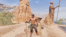 In-game scene from Conan Exiles, depicting a shirtless male character standing on a rocky outcrop in a sunlit desert, surrounded by palm trees and large statues. The environment showcases the game's vast and immersive survival world. Purchase Conan Exiles PC Steam key now at RushGame.co
