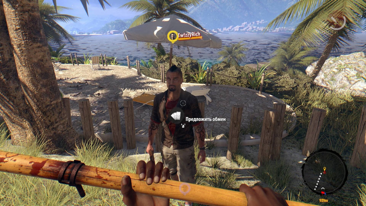  A first-person gameplay shot from Dead Island: Definitive Collection showing a player holding a bloodstained wooden staff while interacting with an NPC standing under an umbrella near a tropical resort. Explore the open world and fight for survival—available now at RushGame.co