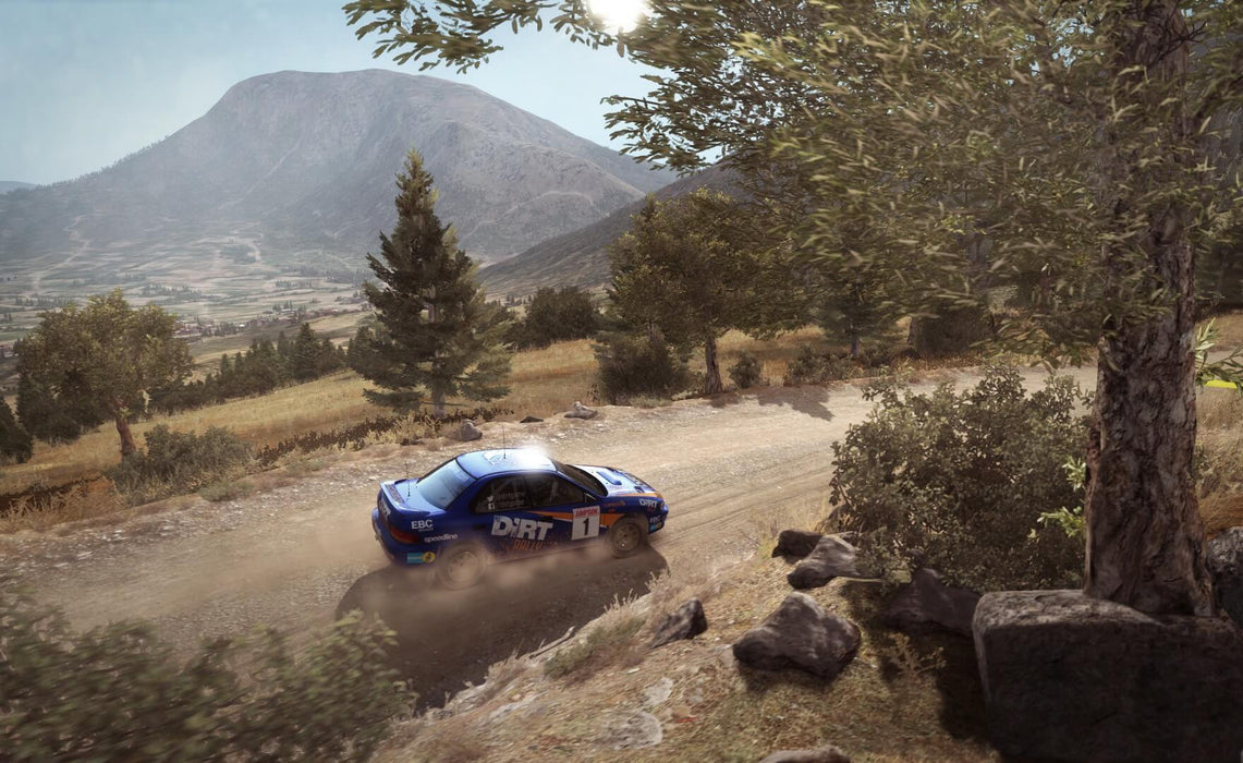 DiRT Rally Gameplay screen – Mountain Rally Track – A blue rally car speeds along a dusty gravel road in a scenic mountain landscape, showcasing DiRT Rally's realistic physics and environmental details. Buy your Steam key now at RushGame.co
