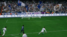 In-game moment from EA Sports FC 24 Ultimate Edition on Default Title & Xbox Series X/S, featuring a powerful shot from a Real Madrid player in a UEFA Champions League match. The goalkeeper dives in an attempt to stop the ball, while the crowd waves white and blue flags in support. Get your EA Sports FC 24 Ultimate Edition CD Key at RushGame.co for instant play