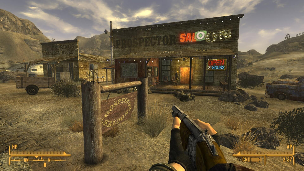 Gameplay screenshot - A first-person view of the player holding a firearm outside the Prospector Saloon in Goodsprings, a rustic desert town in Fallout: New Vegas. Begin your journey through the wasteland with Fallout: New Vegas Ultimate Edition PC Steam CD Key, available for instant download at RushGame.co