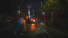 Gameplay from Resident Evil 3 showing Jill Valentine navigating a destroyed city street at night as distant fires rage. Get your Resident Evil 3 Steam key now at RushGame.co and survive Raccoon City's downfall